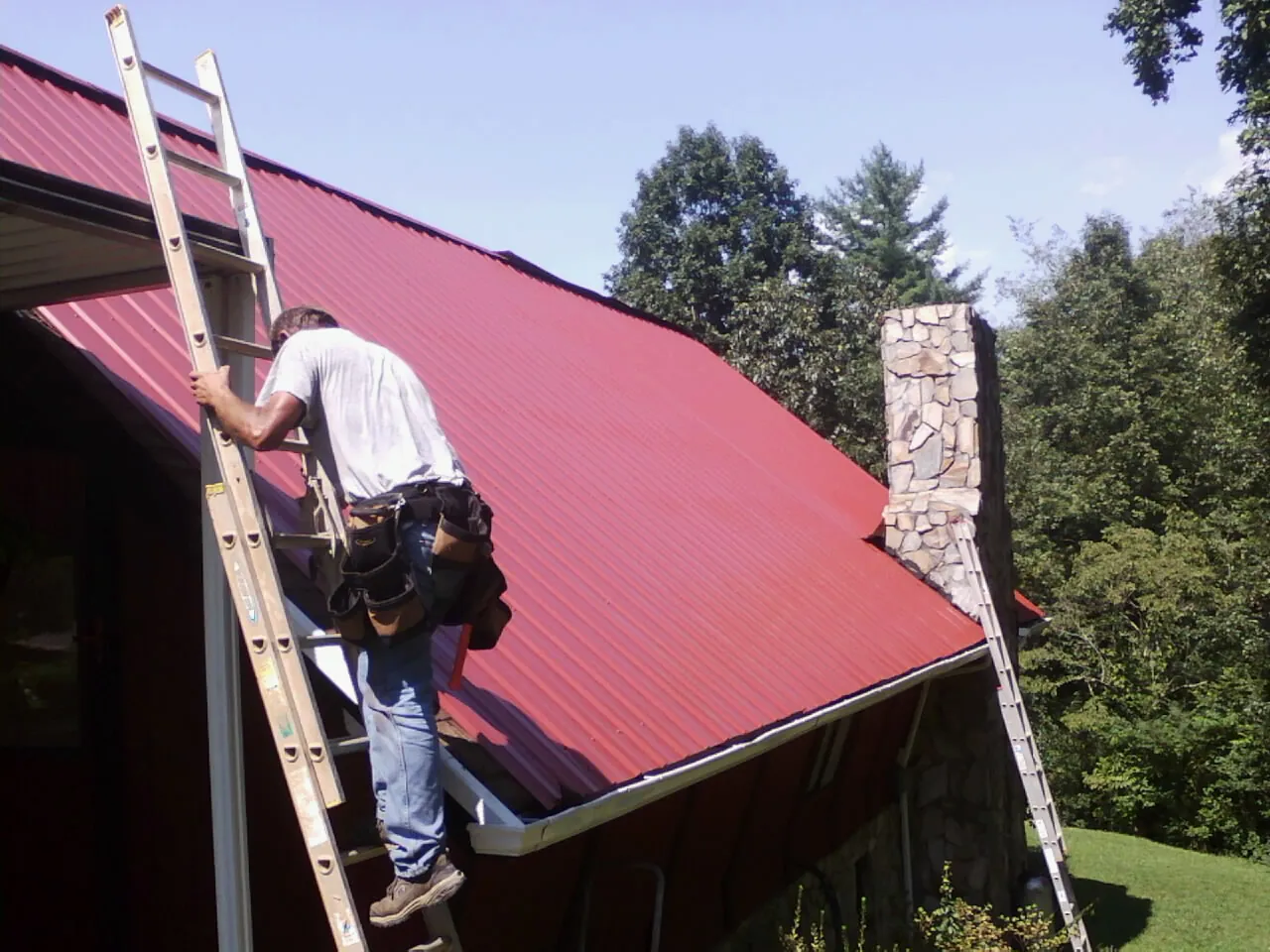 Roofer on a ladder beside a red metal roof and stone chimney — Soffit & Fascia Repair by Roofing Brothers in Ipswich